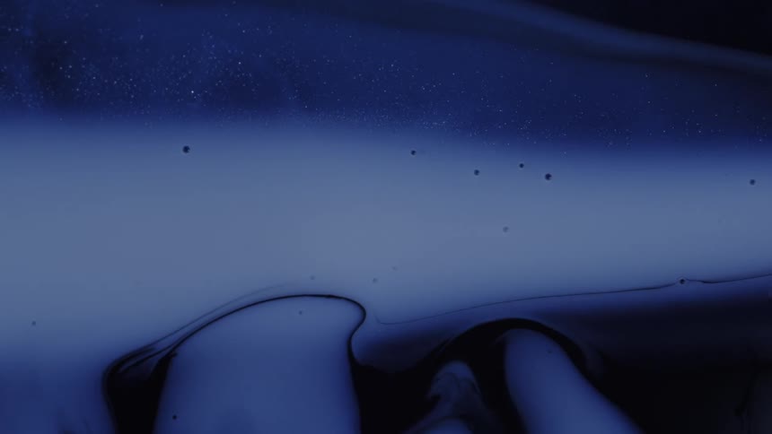 Dark blue and black liquids merge and flow across a surface creating swirling abstract patterns mixed with small glittering particles and the viscous fluids move slowly to form organic shapes and layered textures against a deep dark background