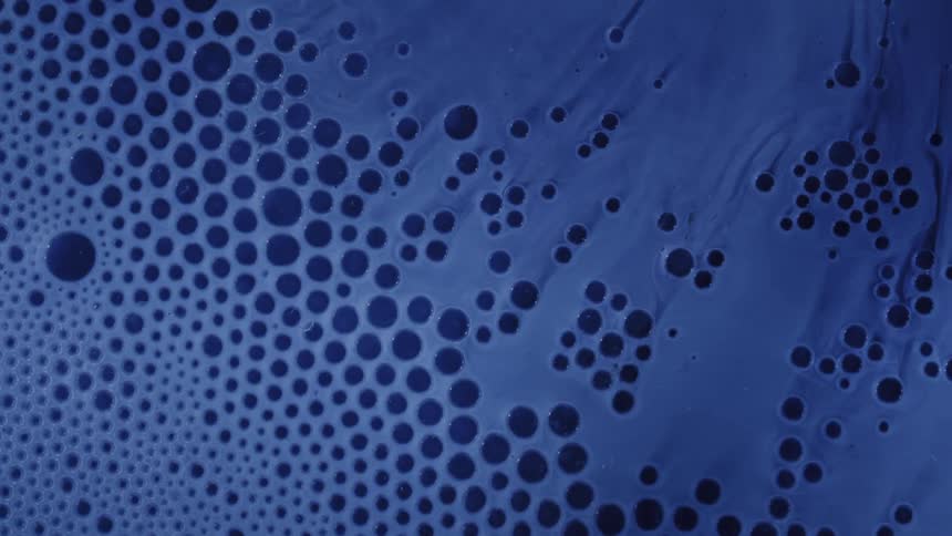 Tiny dark bubbles and deep blue ink spread slowly across a bright blue surface to create a detailed organic pattern