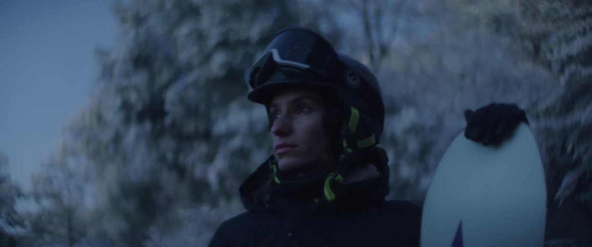 A focused snowboarder in a helmet and winter jacket gazes into the distance while holding a snowboard upright at their side framed by frostcovered trees and cool blue light that emphasizes the stillness and resolve of the cold mountain environment