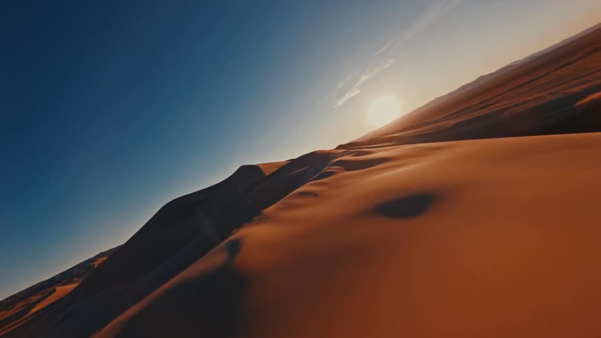 Racing at high speed just inches above the sharp crest of a sand dune the camera captures the vast desert landscape bathed in the warm golden glow of dusk