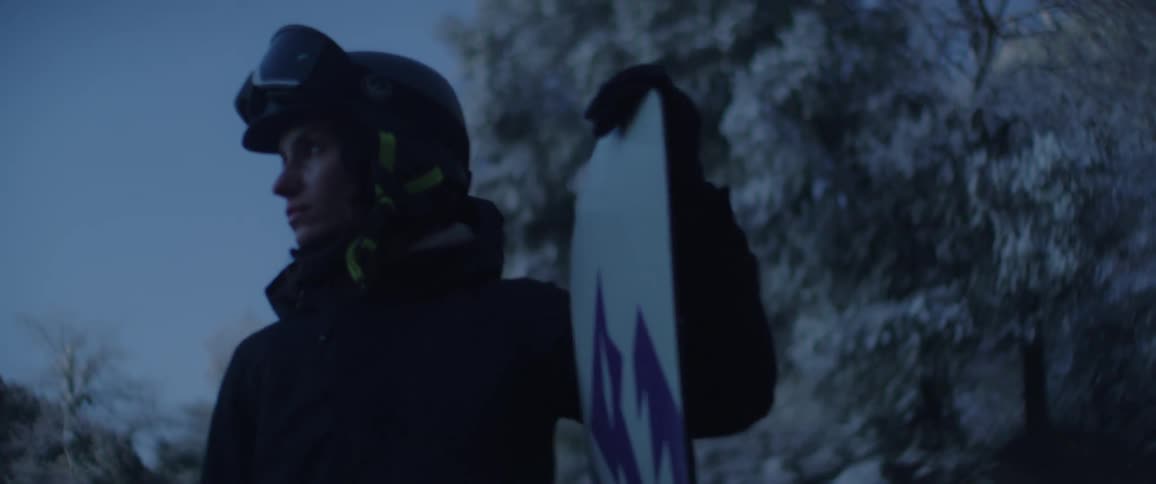 A snowboarder in full winter gear stands in profile holding a snowboard upright at their side silhouetted against frostladen trees and a cold blue sky capturing a calm reflective pause in a wintry mountain setting