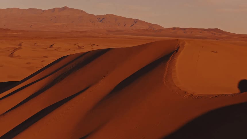Sunlight shines on sand dunes in the desert with mountains in the distance