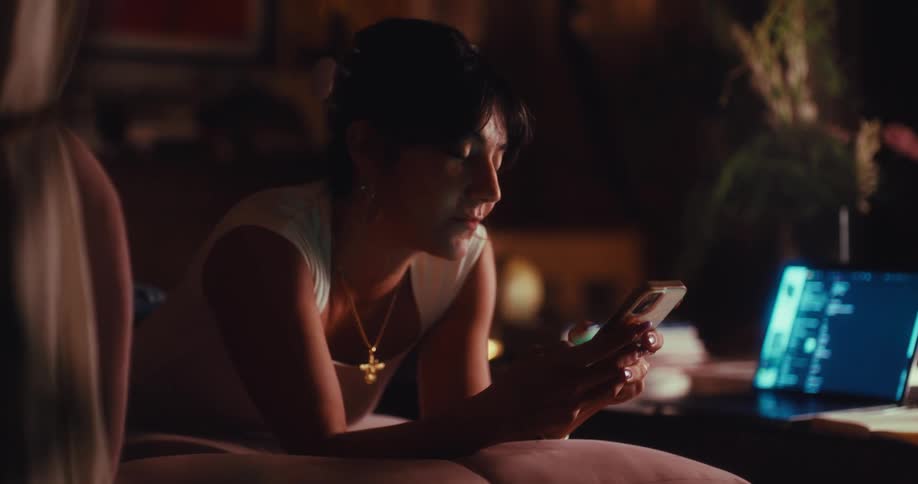 Woman lies next to a table with a laptop computer as she uses her phone