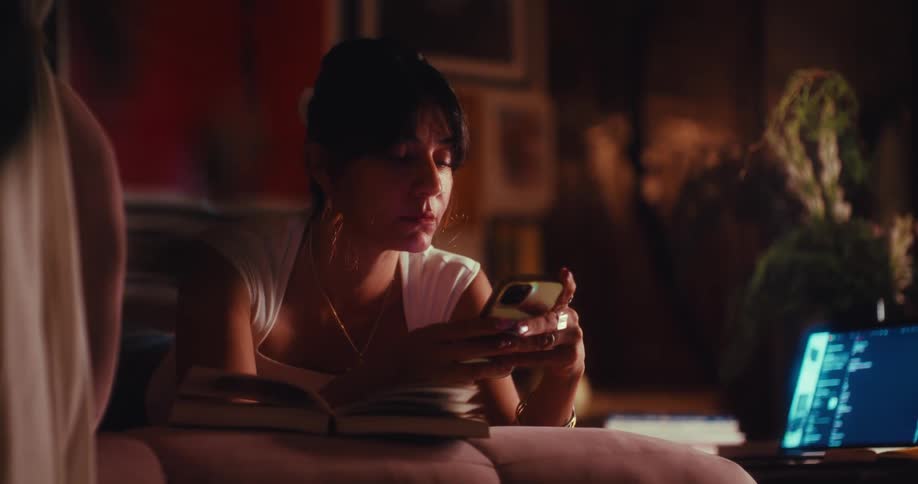 A woman lies on her stomach indoors transitioning from reading an open book to picking up and scrolling through her smartphone
