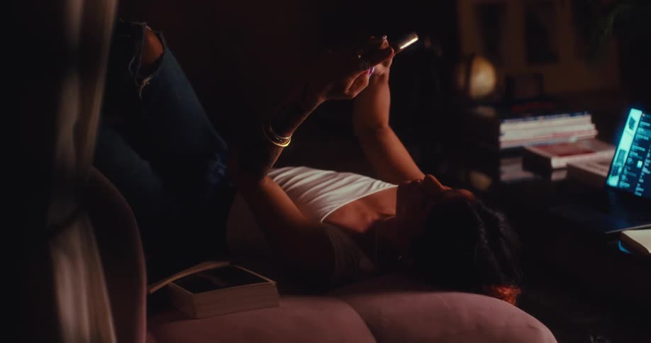 A woman in a white tank top and ripped jeans lies on a pink sofa in a dimly lit room at night scrolling through her phone while a laptop screen glows on a desk nearby surrounded by books and a globe