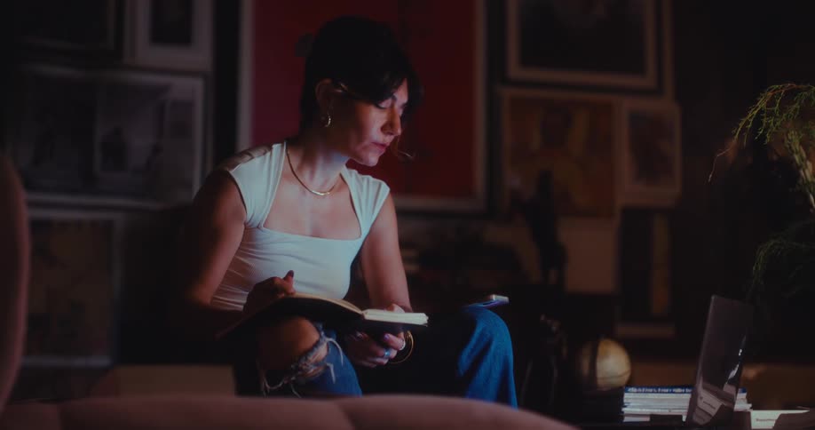 A woman sits in a dark room with warm lighting writing in a notebook on her lap before pausing to rest a pen against her chin while looking thoughtfully off camera