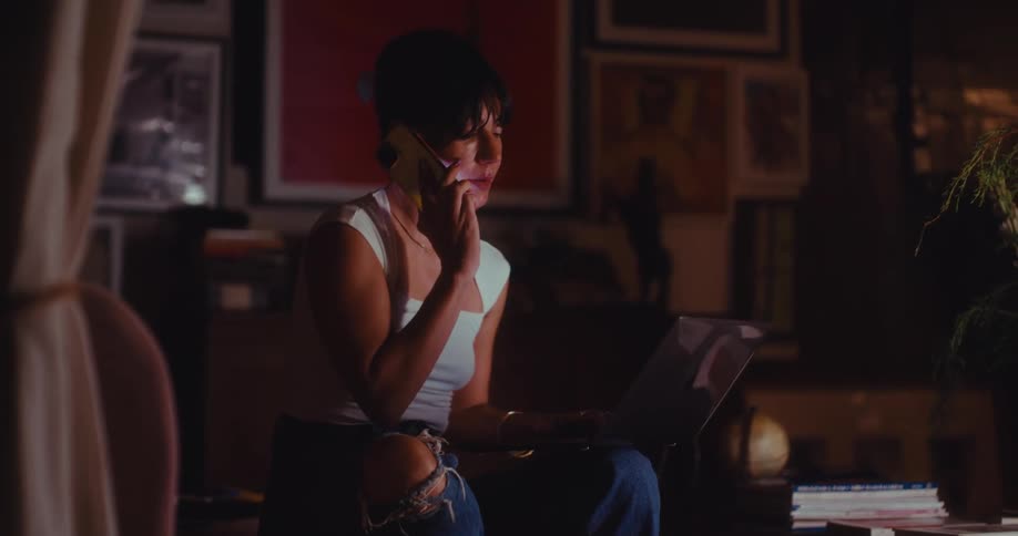 A woman working on her laptop while talking on the phone in a dimly lit living room