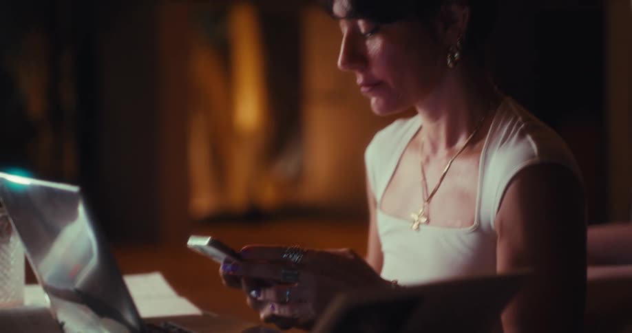 Woman sitting at a table typing on a laptop computer as she stops to use her phone