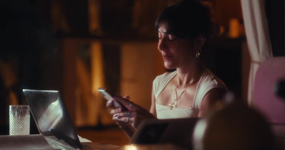 A woman is sitting at a table with a laptop and looking at her phone