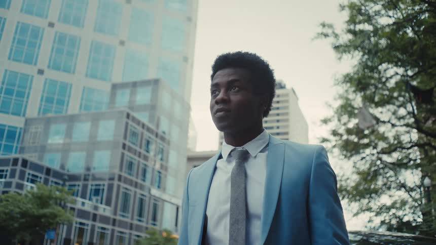 Young adult black man wearing a blue suit and a necktie standing on a street with trees and high rise buildings on the background outdoors during day time