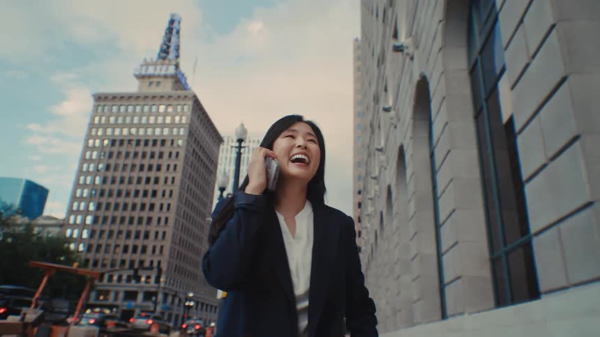 A businesswoman talks on her phone while walking along a bustling city street lined with tall buildings