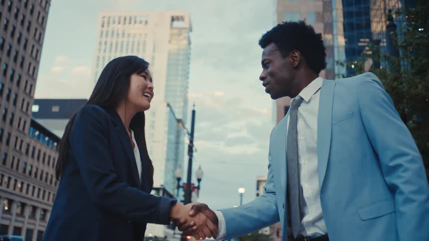 A man in a light blue suit and a woman in a black blazer smile and shake hands while standing on a city street corner surrounded by tall office buildings during the day