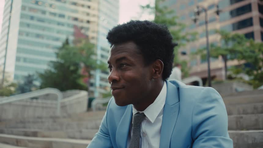 A young black man in a light blue suit sits on outdoor stone stairs in a city looking offcamera with a slight smile while a person walks by in the background near tall office buildings and trees