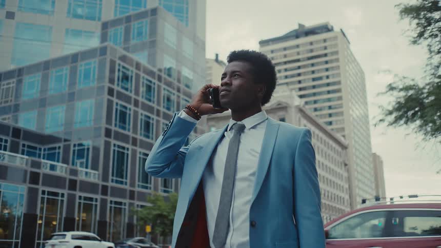 Young adult black man wearing a blue suit and necktie walking on a metropolitan area while talking on the phone outdoors during day time