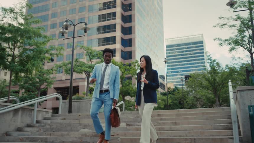 Man and woman walk and talk in an urban area with tall buildings and surrounding trees