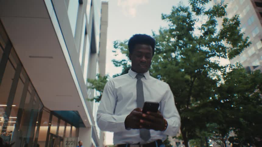 Man uses a smartphone while walking along an urban sidewalk beside buildings and trees