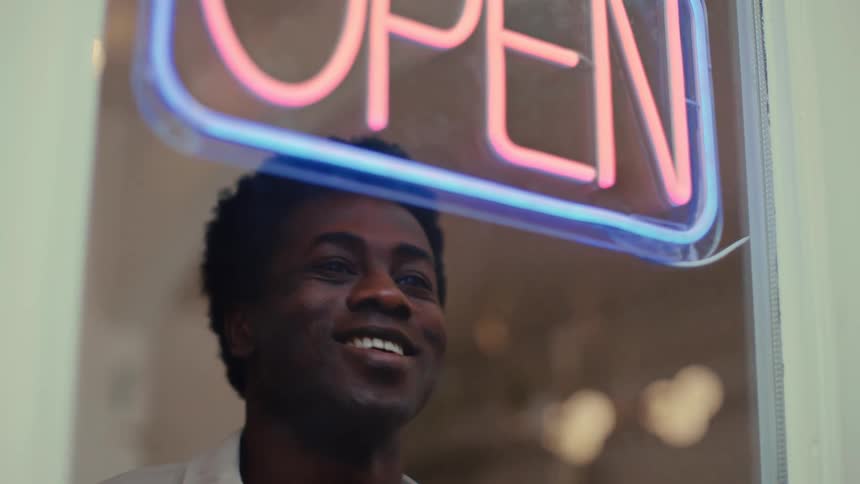 A man smiles warmly behind a neon open sign in a shop window