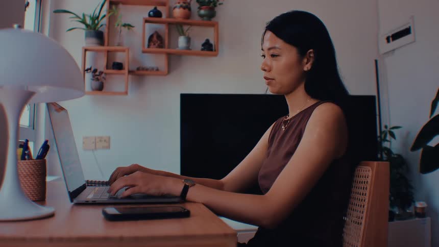 A woman with long black hair wearing a brown sleeveless top sits at a wooden desk and types on a laptop computer in a bright room with potted plants