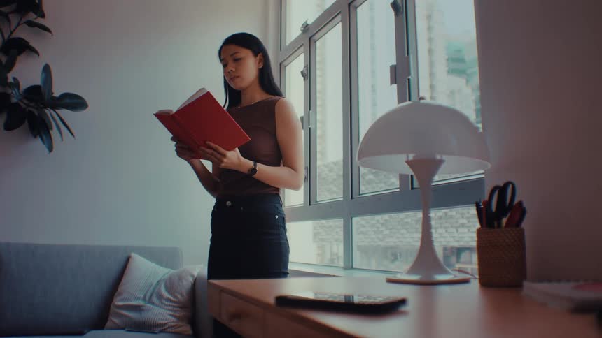 A woman with long black hair wearing a brown sleeveless top stands by a window in a bright room holding a red book before reaching for a smartphone on a wooden desk