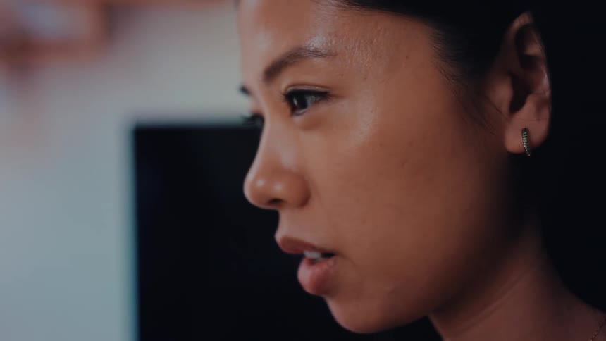 An asian woman in closeup profile with a pensive expression with a lcd tv in the back on a dimly lit indoor environment