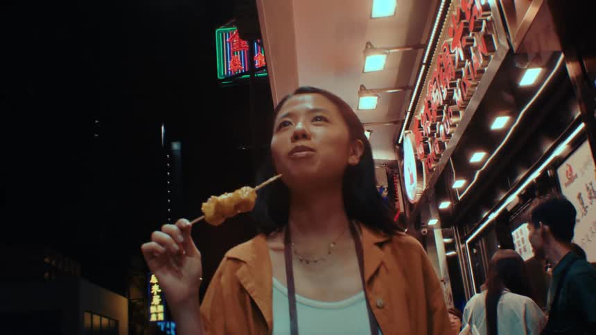 A young woman walks through a brightly lit urban night market while eating food from a wooden skewer and other people are in the background