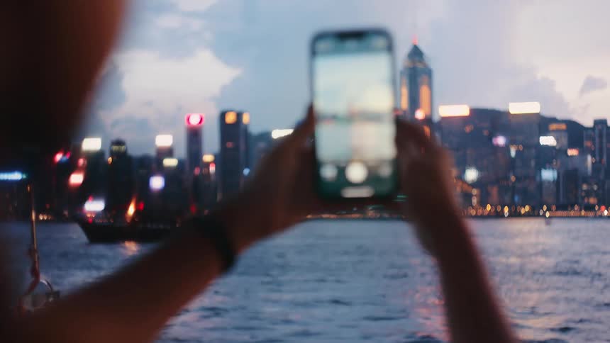 A woman uses a smartphone to photograph a illuminated city skyline across a waterfront at dusk as a traditional junk boat sails past the brightly lit skyscrapers