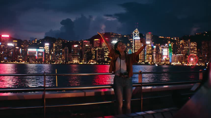 A woman takes a selfie by the waterfront at night with the glowing city skyline in the background