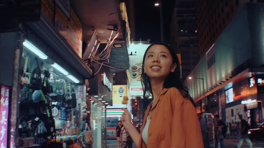 A young woman walking along a busy street at night holding a piece of food on a stick and taking happy bites as she enjoys the lively atmosphere around her