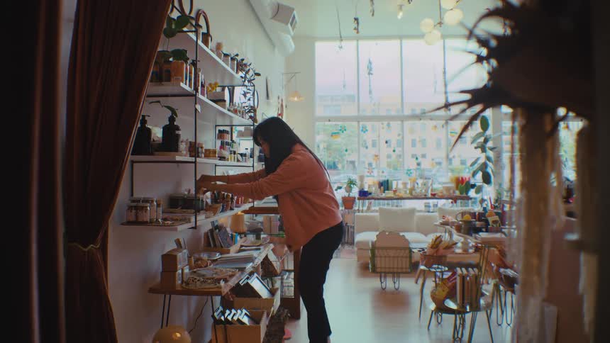 A young woman wearing a brown jacket sorting products inside a retail shop at midday organizing items neatly on the shelves as sunlight filters through the storefront windows