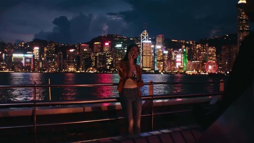 The woman is holding her camera and talking on her cell phone by the big city