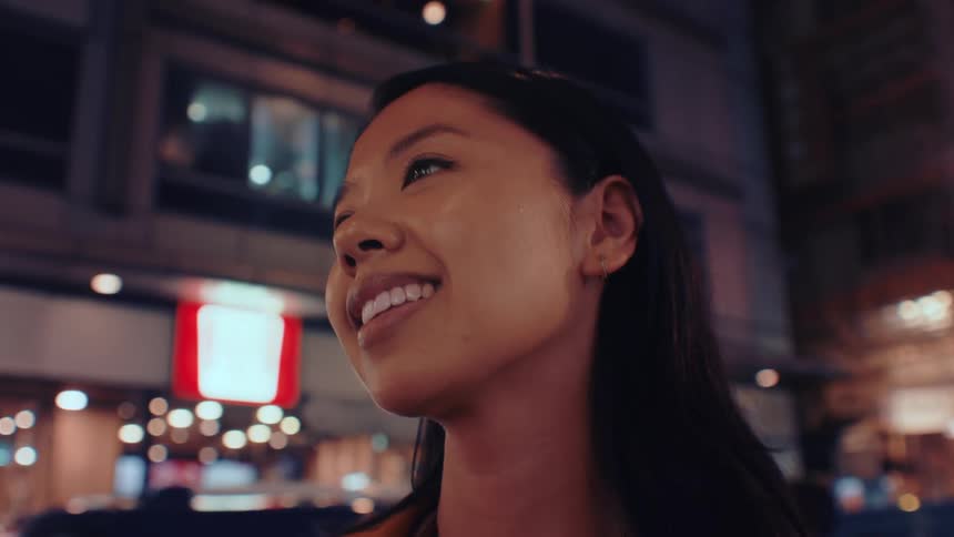 A young asian woman standing on the sidewalk at night looking around with a warm smile as the city lights glow softly around her