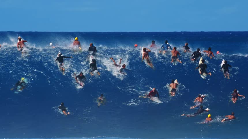 A large crowd of surfers scrambles and paddles through a massive crashing blue wave under a clear skythe churning white foam tosses boards and people in multiple directions as they navigate the powerful ocean energy