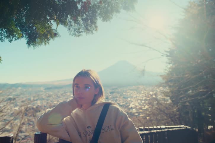 A woman in a tan hoodie sits at a mountain overlook with her hand on her chin looking out over a city toward mountains as the sun shines from the upper right of the frame