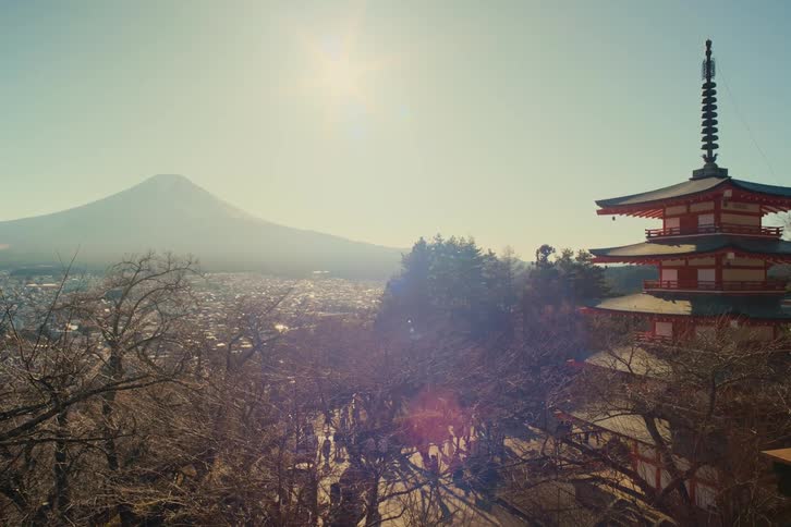 Golden sunlight bathes the iconic chureito pagoda where people walk the grounds with and bare cherry blossom trees offering a breathtaking view of a sprawling city nestled in the valley beneath the majestic hazy peak of mount fuji