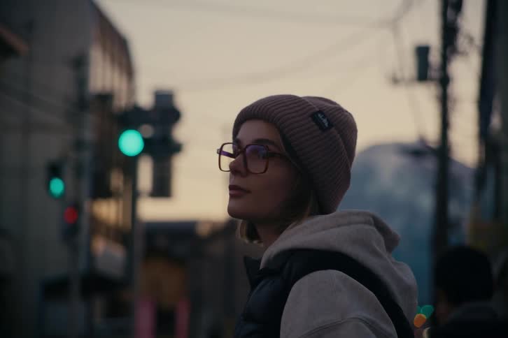 Woman wears a beanie and glasses on an urban street at dusk with a lit traffic signal in the background