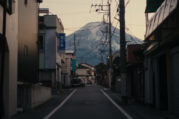 A car drives down a narrow town street lined with utility poles and traditional buildings toward the towering snowcapped peak of mount fuji as evening light casts a soft glow over the landscape
