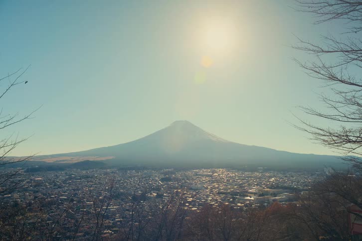High above the valley bright sunlight creates a shimmering lens flare over a vast city layout that stretches toward the base of mount fuji standing as a silent majestic backdrop under a clear blue sky
