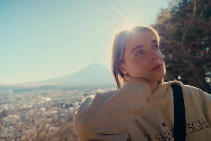 A young woman in a light gray hoodie stands outdoors on a hillside overlooking a sprawling city softly backlit by warm sunlight that creates a gentle lens flare as she gazes thoughtfully in the distance