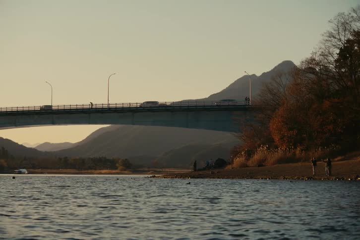 Bridge spans a river with moving vehicles and people along the shore in front of mountains at dusk