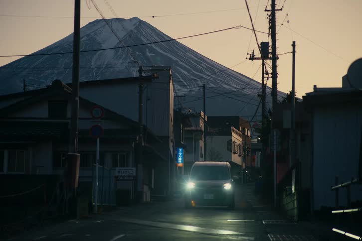 Urban street at dusk frames mount fuji in the background with an approaching vehicle and aligned power poles