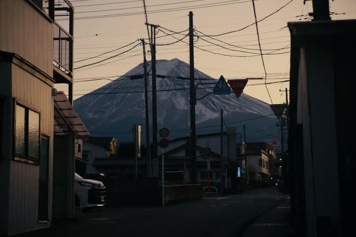 A quiet japanese street lined with utility poles and houses leads the eye toward the massive snowdusted silhouette of mount fuji towering over the neighborhood which is partially silhouetted by the soft fading evening light