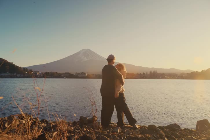 A couple embraces by a lakeside at sunset with a snow capped mountain in the background