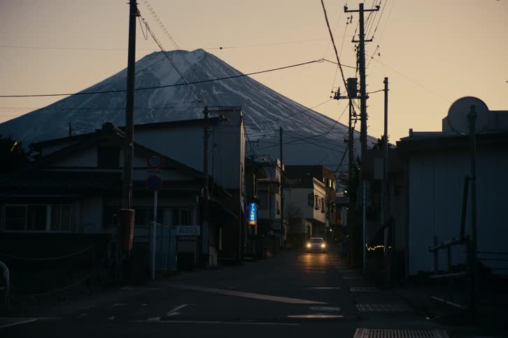 A stationary shot of a narrow street at dusk with mount fuji looming in the background while cars drive past in the foreground and down the road