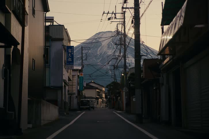 A quiet narrow street lined with small buildings and shuttered storefronts leads toward a snowdusted mountain rising dramatically in the background framed by a dense web of power lines and poles under warm fading light