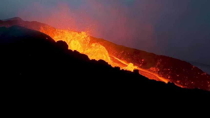 A violent lava fountain erupts from a glowing volcanic channel hurling incandescent molten rock into the air as thick steam and smoke rise around the blazing flow illuminating the rugged terrain