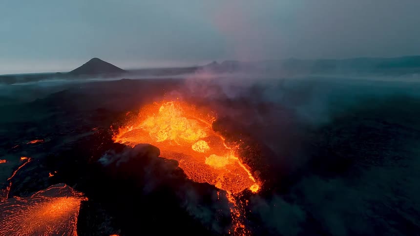 A drone captures an aerial view of an active volcano at dusk as molten lava bubbles inside a crater and flows across a dark rocky landscape while thick plumes of smoke rise into the dim sky