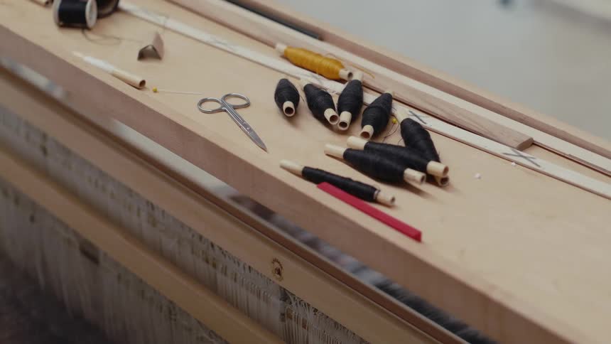 A collection of black and yellow thread spools metal scissors and thin wooden rods rests neatly on the wooden frame of a traditional weaving loom as fine white threads stretch across the background