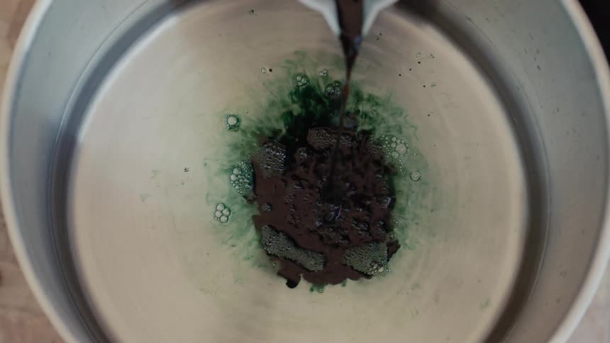 As a stream of dark green concentrated ink is poured into a large metal vat of clear water it blossoms into a vibrant emerald cloud that rhythmically expands and swirls across the shimmering surface