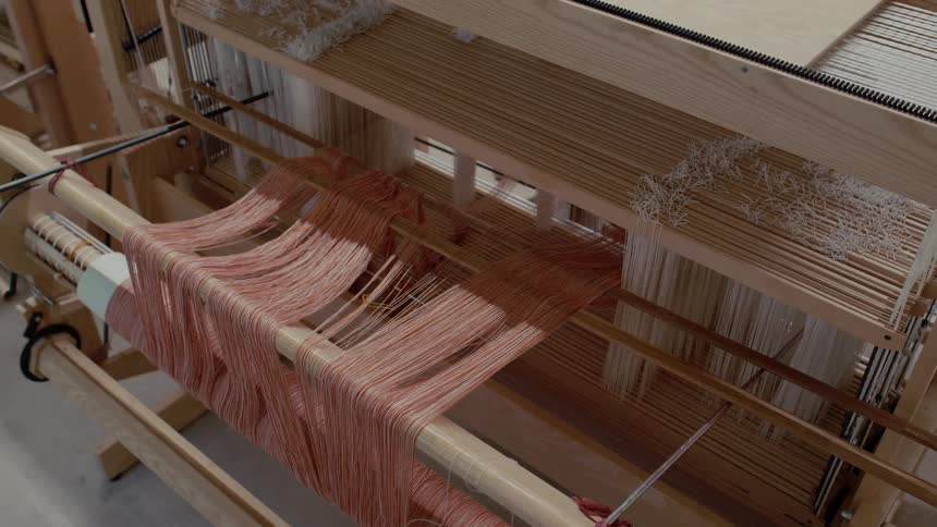 Intricate peachcolored threads are meticulously arranged across the wooden frame of a traditional weaving loom highlighting the complex mechanical beauty of textile preparation in a quiet studio