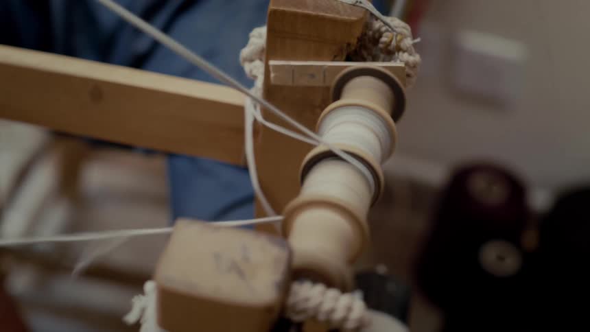 A handcrafted wooden spindle mounted on a simple frame with multiple taut white threads wound and stretched around its smooth cylindrical surface